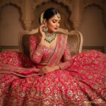 Hot Pink Rani Pink lehenga choli set with heavy gold zari and shisha mirror work worn by a model in a traditional seated pose. Styled with an elaborate Polki and emerald choker necklace, jhumkas, maang tikka, and gajra-adorned bun.
